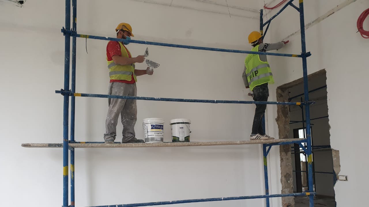 Two men stand on scaffolding in a room and paint a wall white.