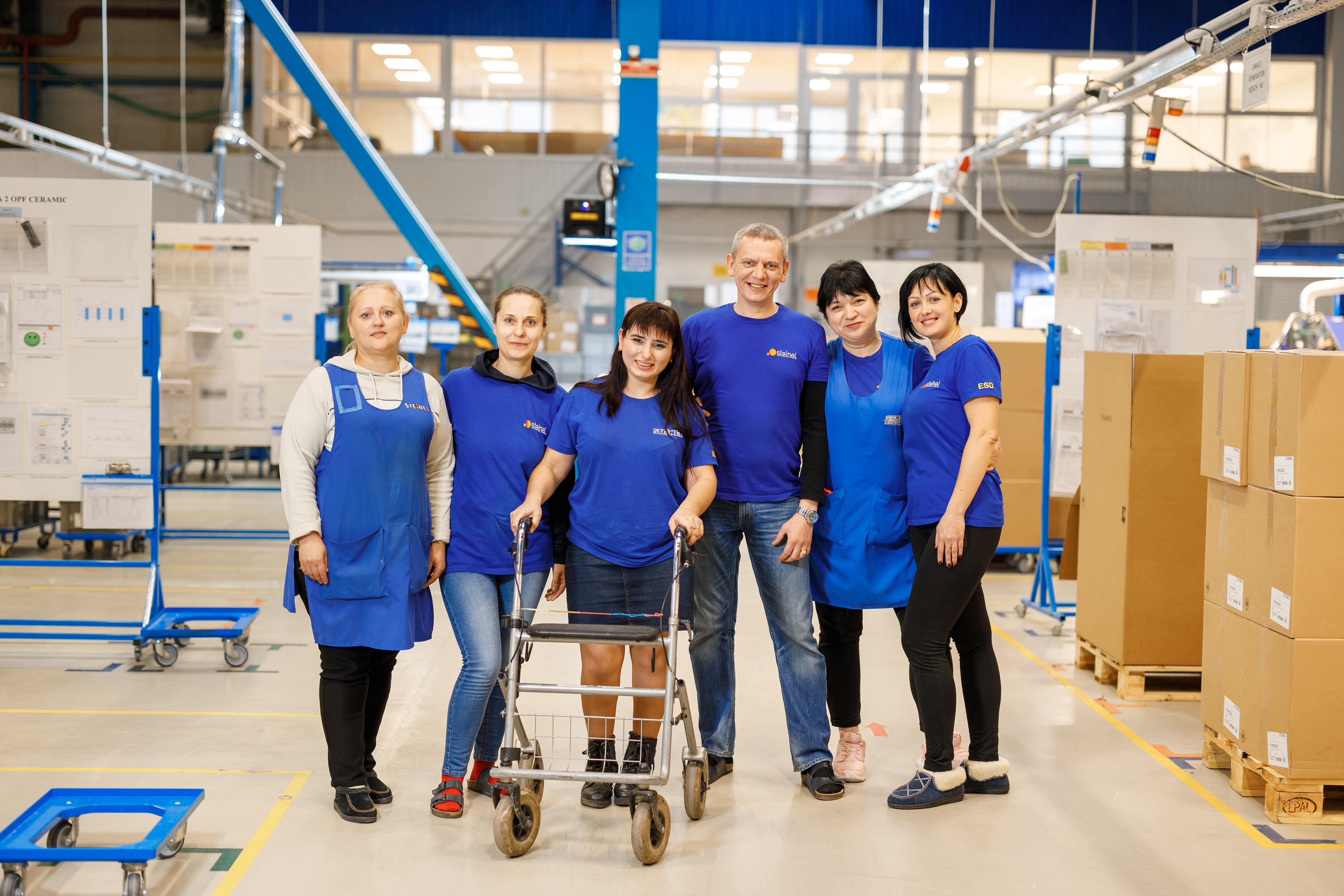 Elena stands with her walker with colleagues. They all wear the same blue, staff t-shirt and smile for the camera.