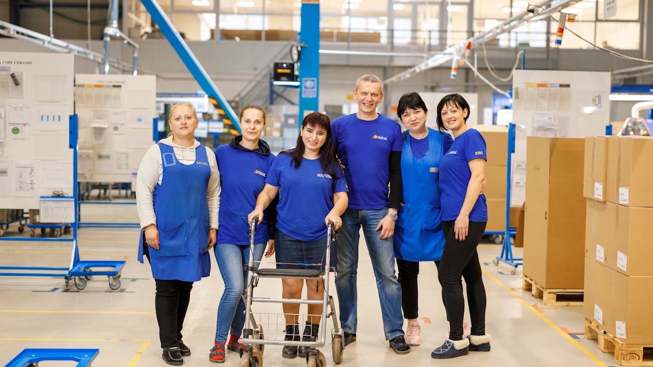Elena stands with her walker with colleagues. They all wear the same blue, staff t-shirt and smile for the camera.