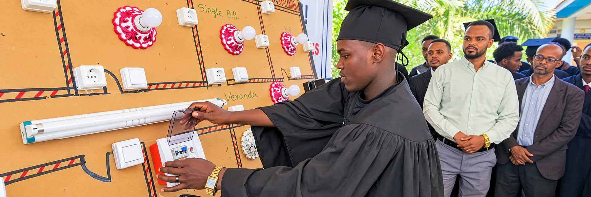 A young man wears a graduation black gown and cap and operates a demonstration electrical panel with other people looking on.