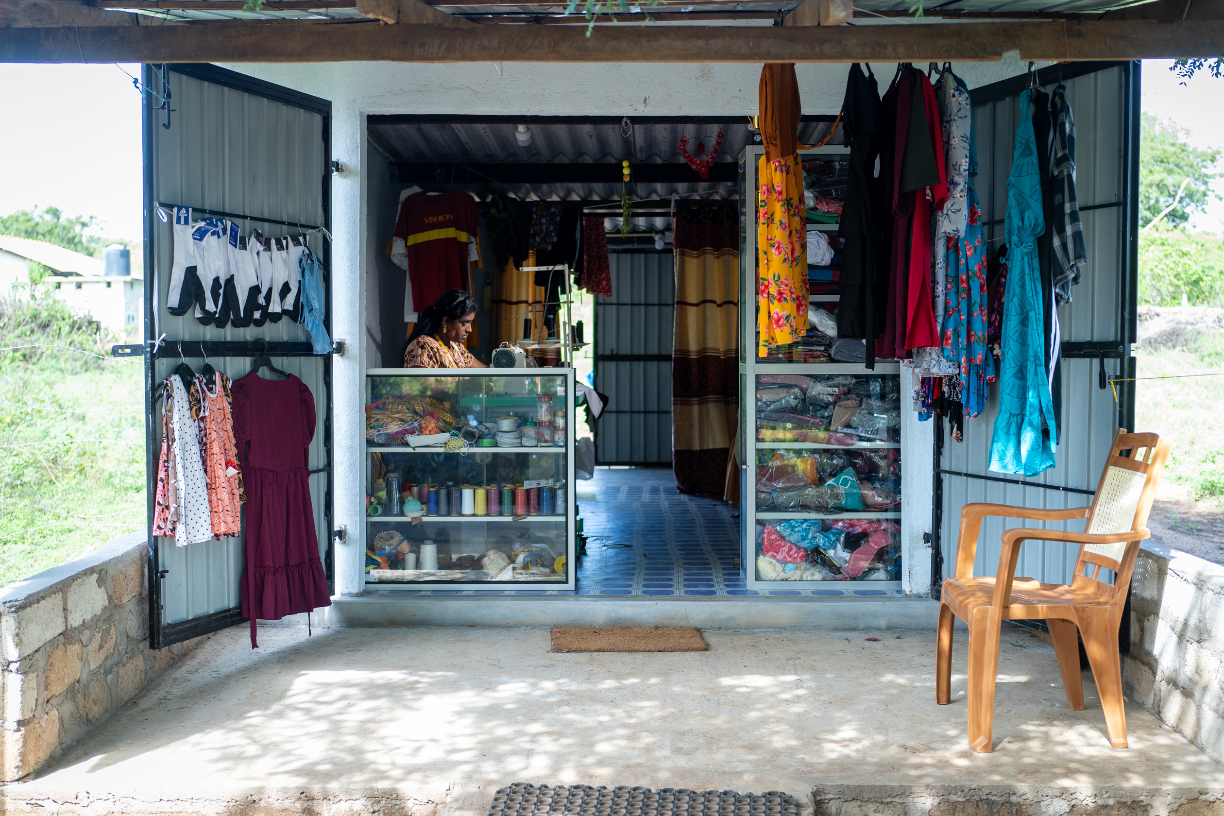 Leena Penildas Thatkuras works at a sewing machine in her tailoring shop. Threads and fabrics are in display cabinets at the front of the shop and garments hang in the foreground.