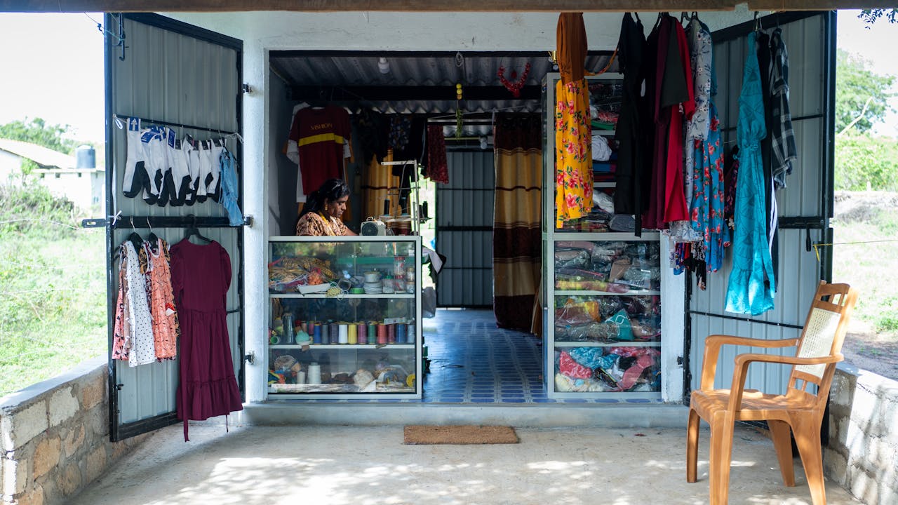 Leena Penildas Thatkuras works at a sewing machine in her tailoring shop. Threads and fabrics are in display cabinets at the front of the shop and garments hang in the foreground.
