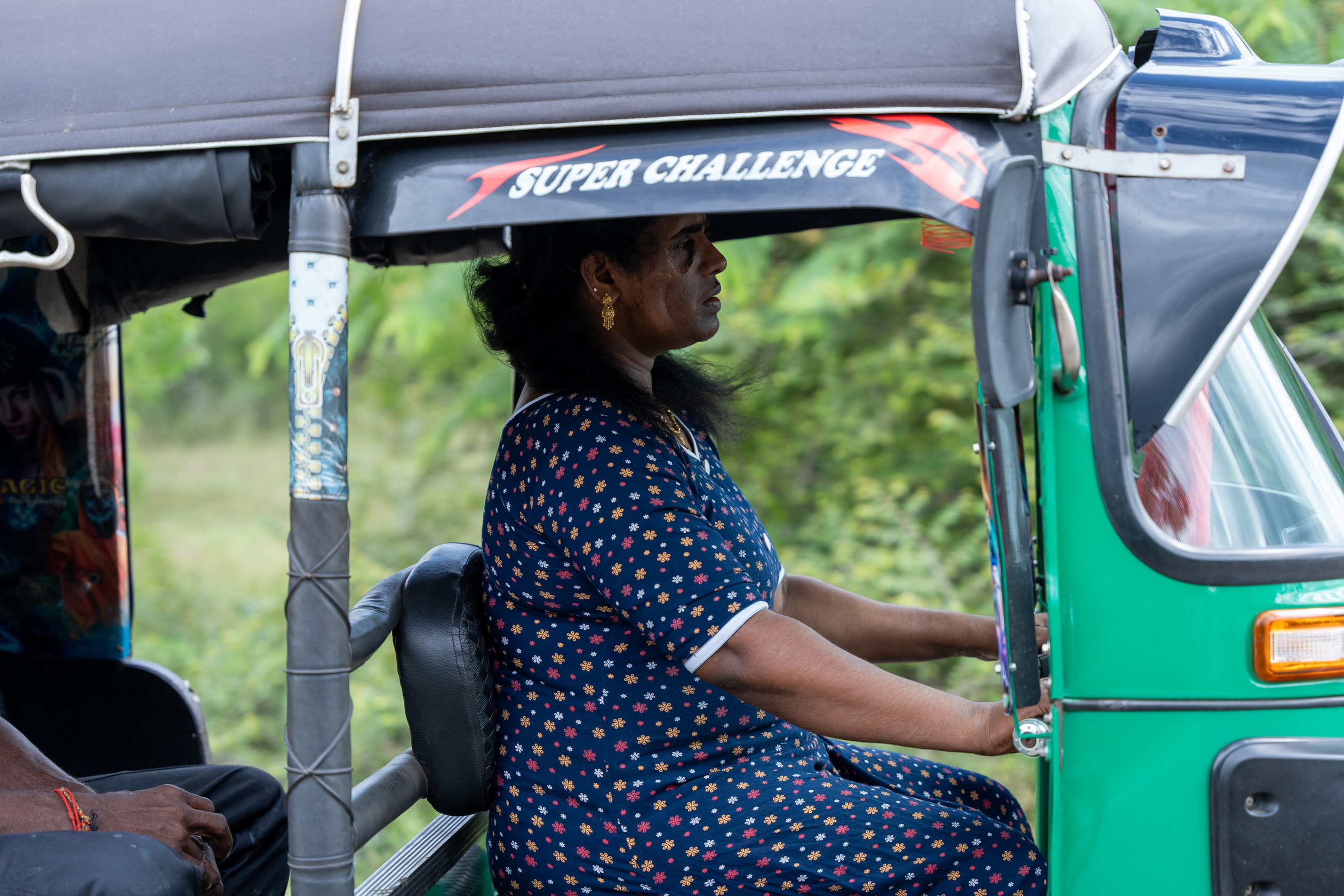 Leena Penildas Thatkuras drives a three-wheeler along a rural road. A passenger sits in the back.