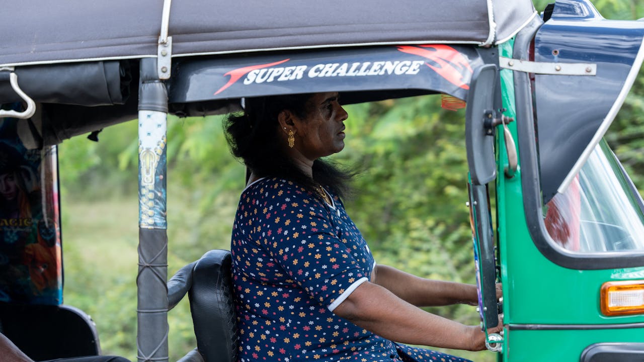 Leena Penildas Thatkuras drives a three-wheeler along a rural road. A passenger sits in the back.