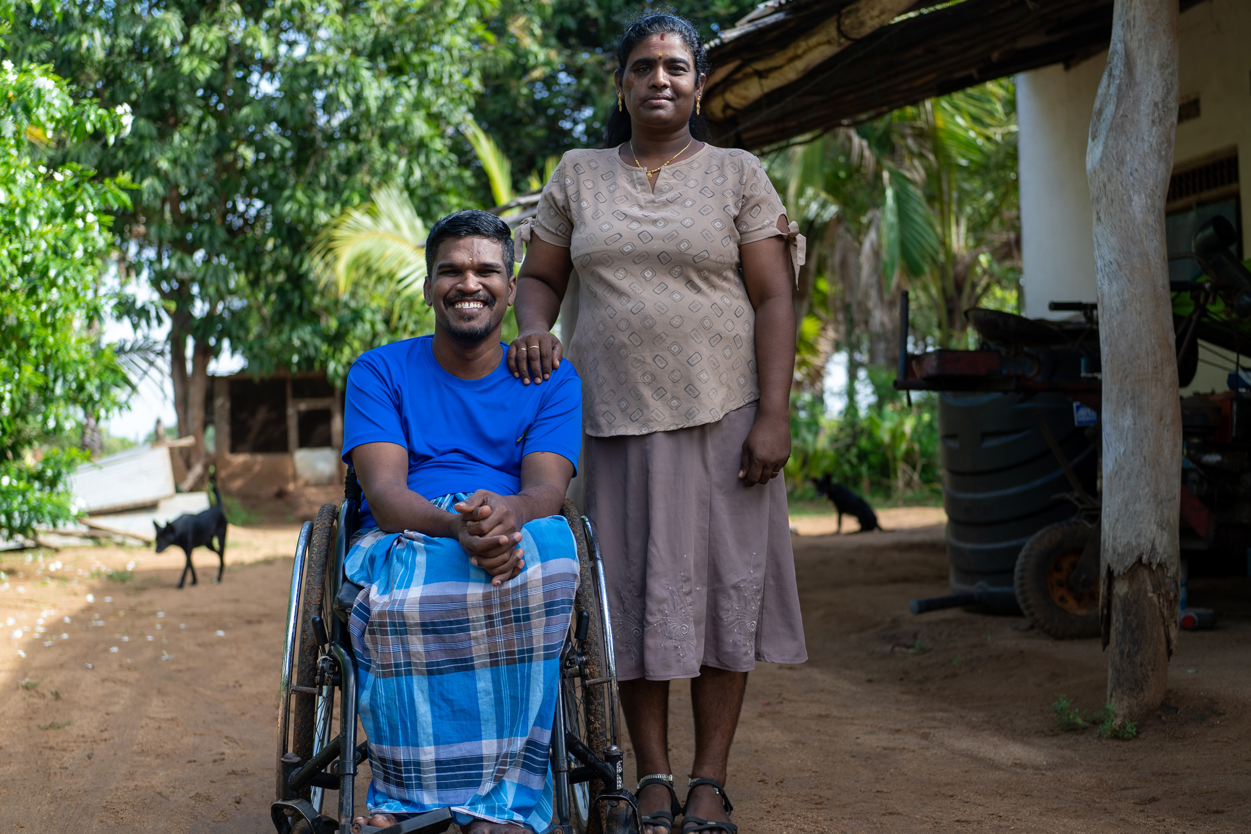 In a garden, Leena Penildas Thatkuras stands beside her husband, with her hand on his shoulder, smiling. Her husband smiles broadly, sitting in a wheelchair.