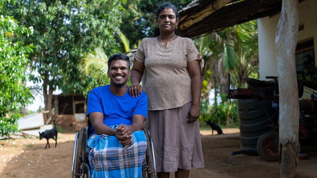 In a garden, Leena Penildas Thatkuras stands beside her husband, with her hand on his shoulder, smiling. Her husband smiles broadly, sitting in a wheelchair.