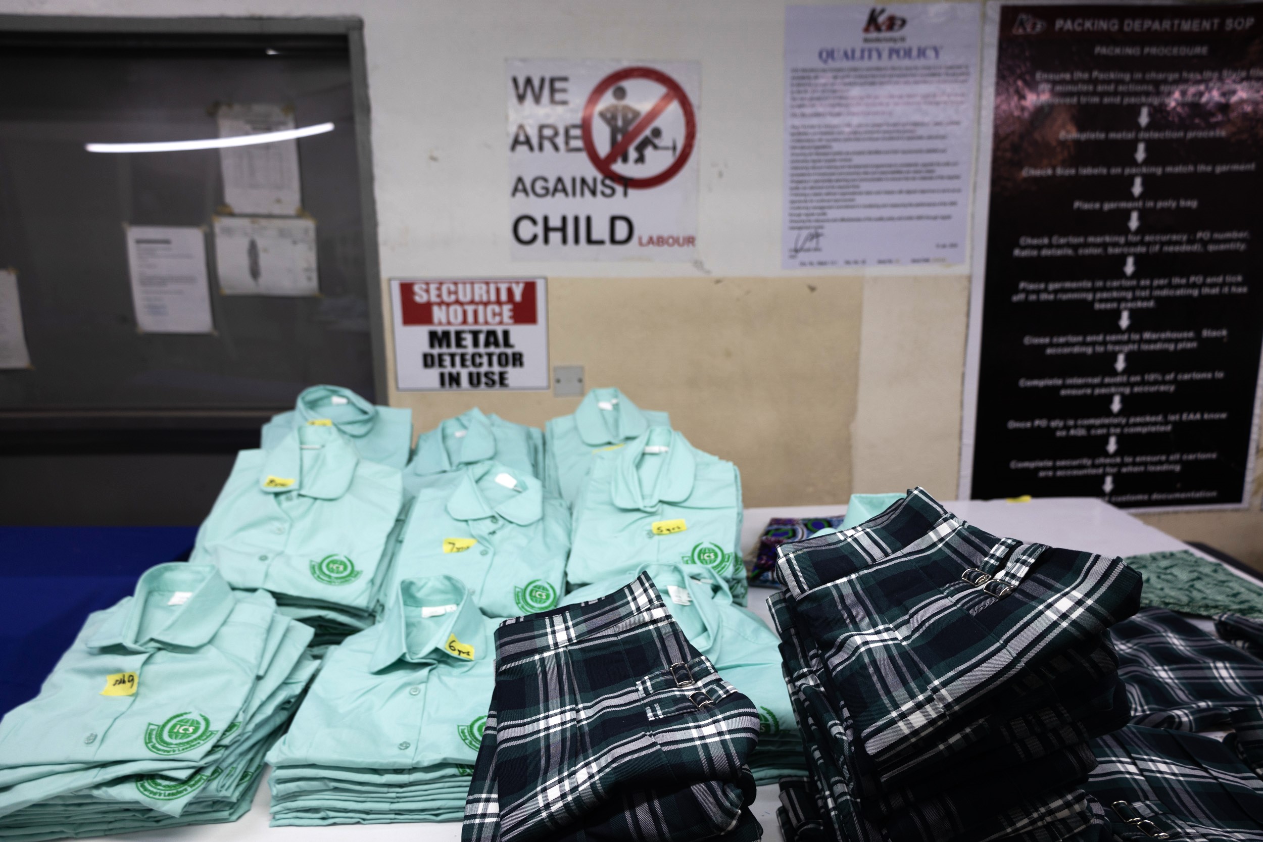 Shirts and skirts are neatly folded on a work surface. In the background, a poster indicates: 'We are against child labour'.