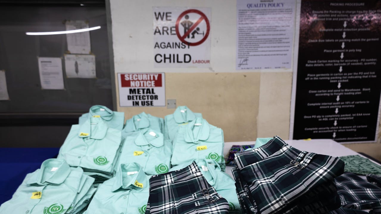 Shirts and skirts are neatly folded on a work surface. In the background, a poster indicates: 'We are against child labour'.