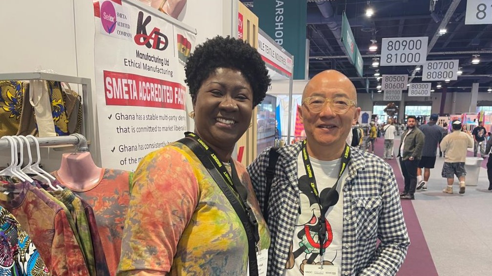 Linda Ampah smiles at the camera with another trade show participant, a man. They stand next to her KAD Manufacturing Limited stall. She wears one of the colourful dresses she is showcasing at her stall.