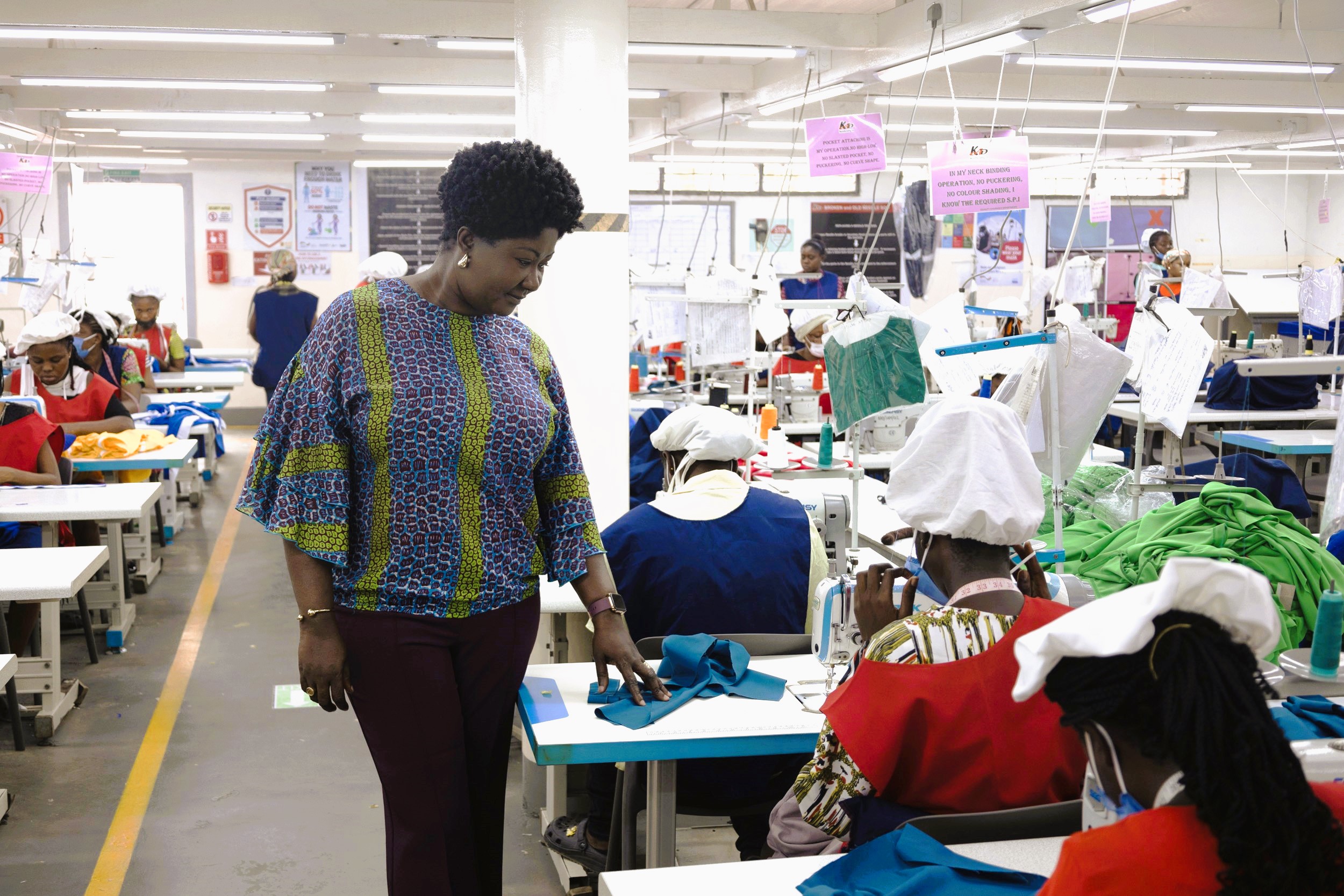 Linda Ampah stands near a woman who is working on a sewing machine in the factory. There are other workers sewing in the background.