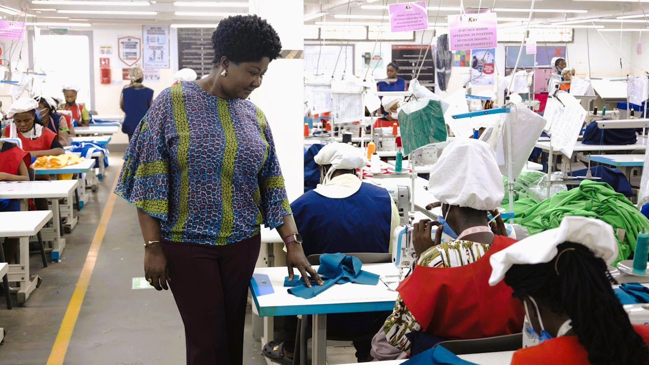 Linda Ampah stands near a woman who is working on a sewing machine in the factory. There are other workers sewing in the background.