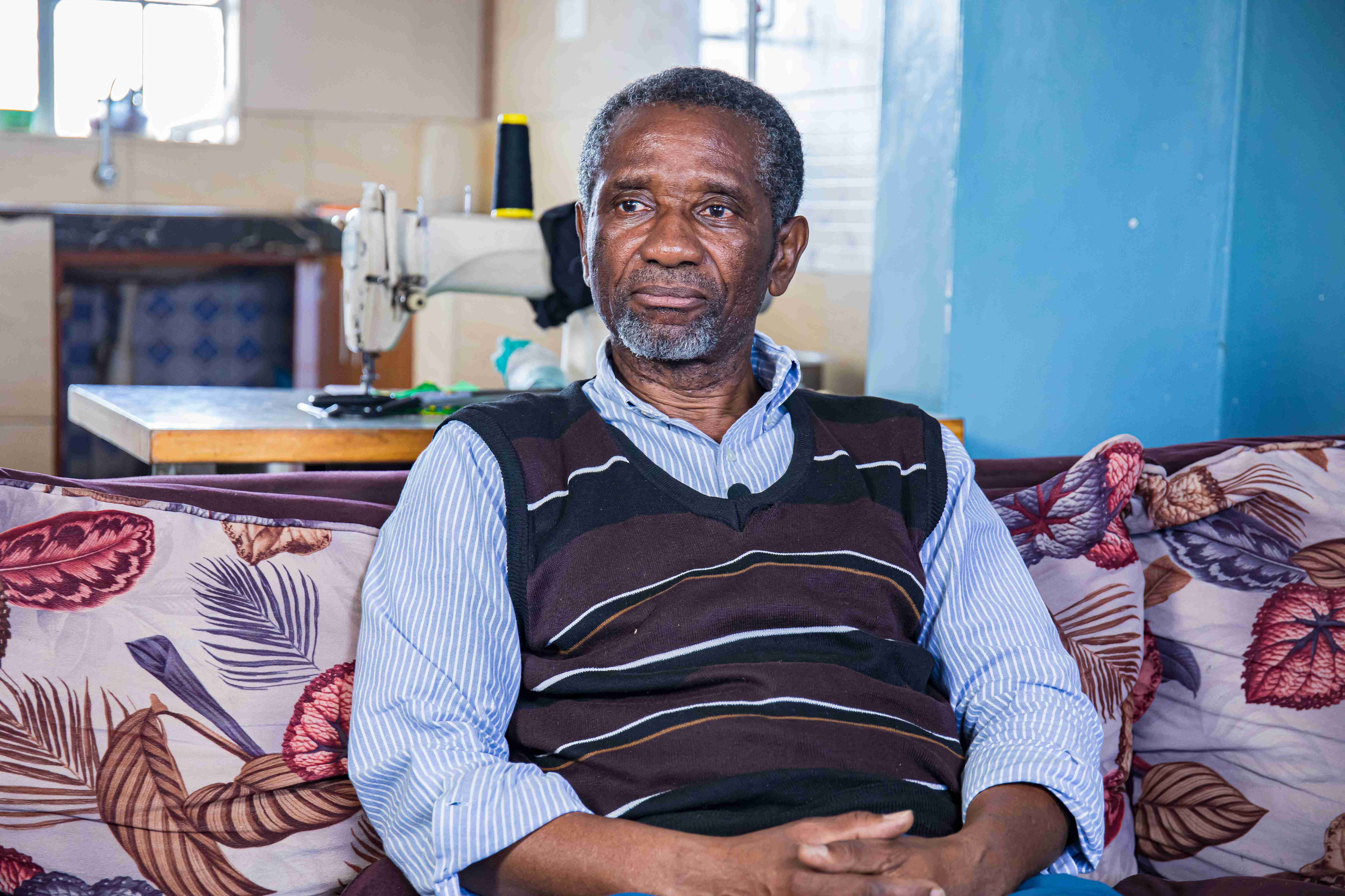 Celestin Mbaruku sits on a sofa, with his hands folded, looking into the distance. Behind him, there is a sewing machine. (Nairobi, Kenya 2024)
