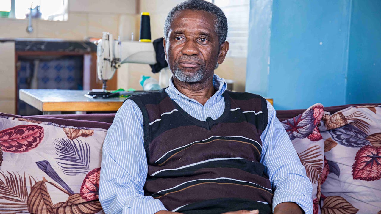 Celestin Mbaruku sits on a sofa, with his hands folded, looking into the distance. Behind him, there is a sewing machine. (Nairobi, Kenya 2024)