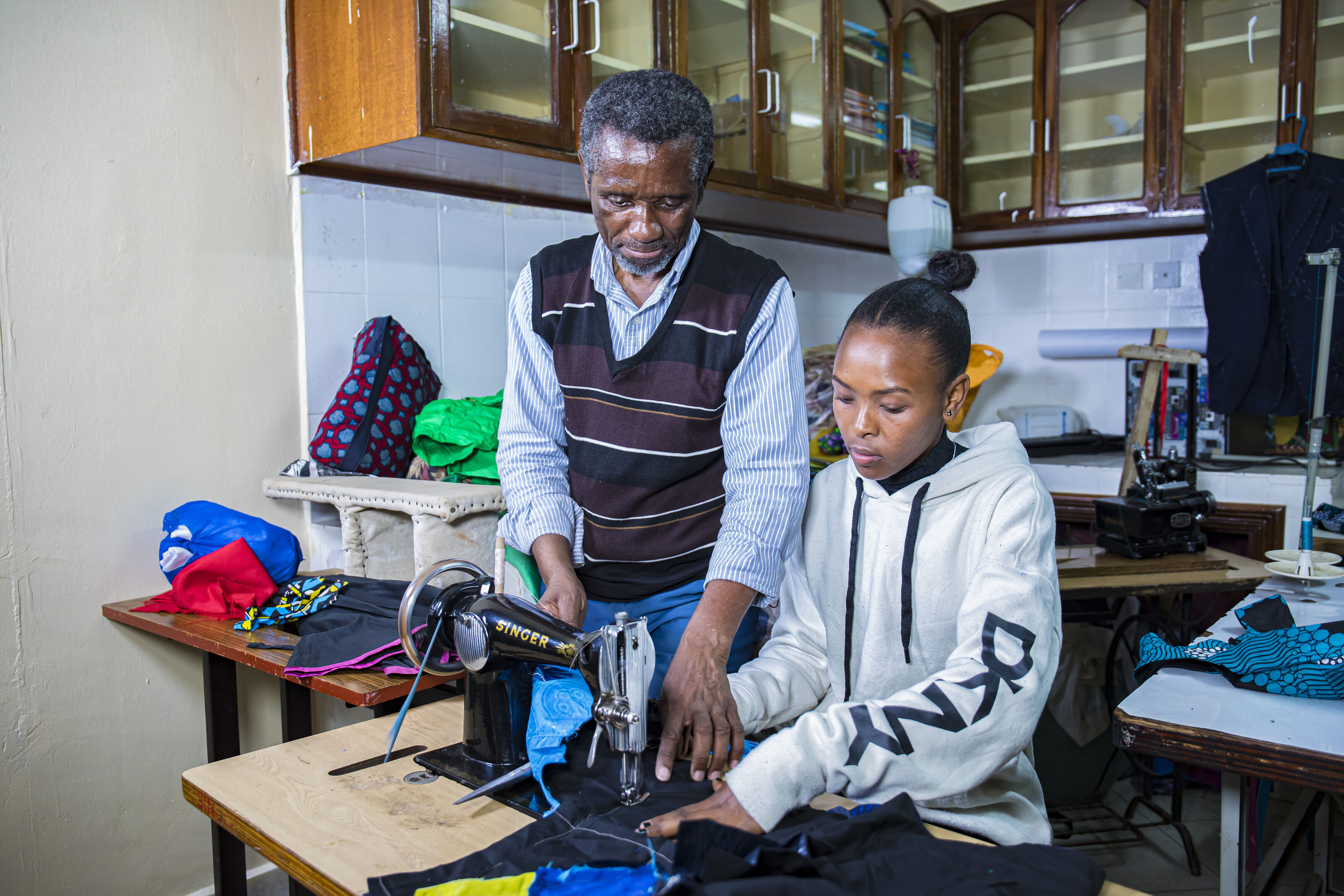 Celestin Mbaruku stands beside a sewing trainee and helps to align the fabric on the sewing machine. (Nairobi, Kenya 2024)