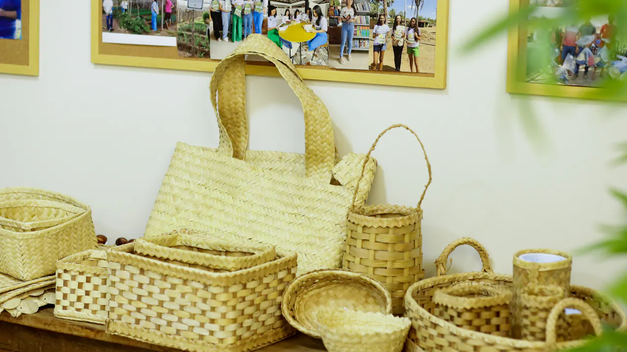Cestas, bolsos y recipientes tejidos están expuesto sobre una mesa. En la pared, se muestran fotos de los grupos de artesanas que elaboraron los productos artesanales.