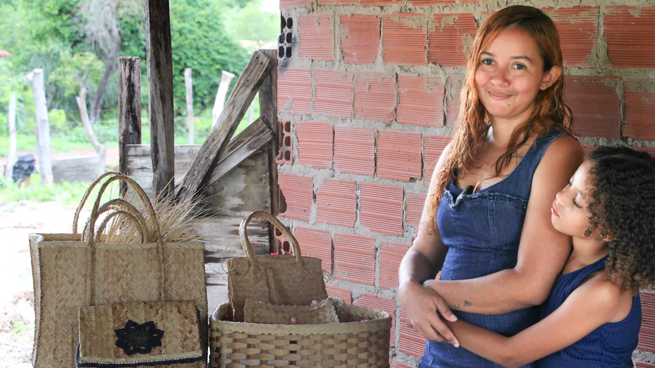 A portrait of Fátima Laine Sousa Santos. She stands smiling with her young daughter beside a table displaying handmade bags and baskets woven from carnauba.