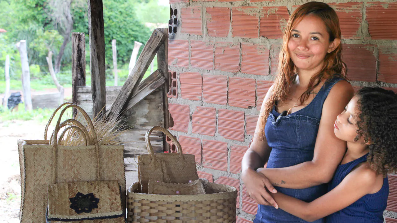 Portrait de Fátima Laine Sousa Santos. On la voit souriante à côté de sa petite fille près d’une table où sont exposés des sacs et des paniers tissés à la main à partir de carnauba.