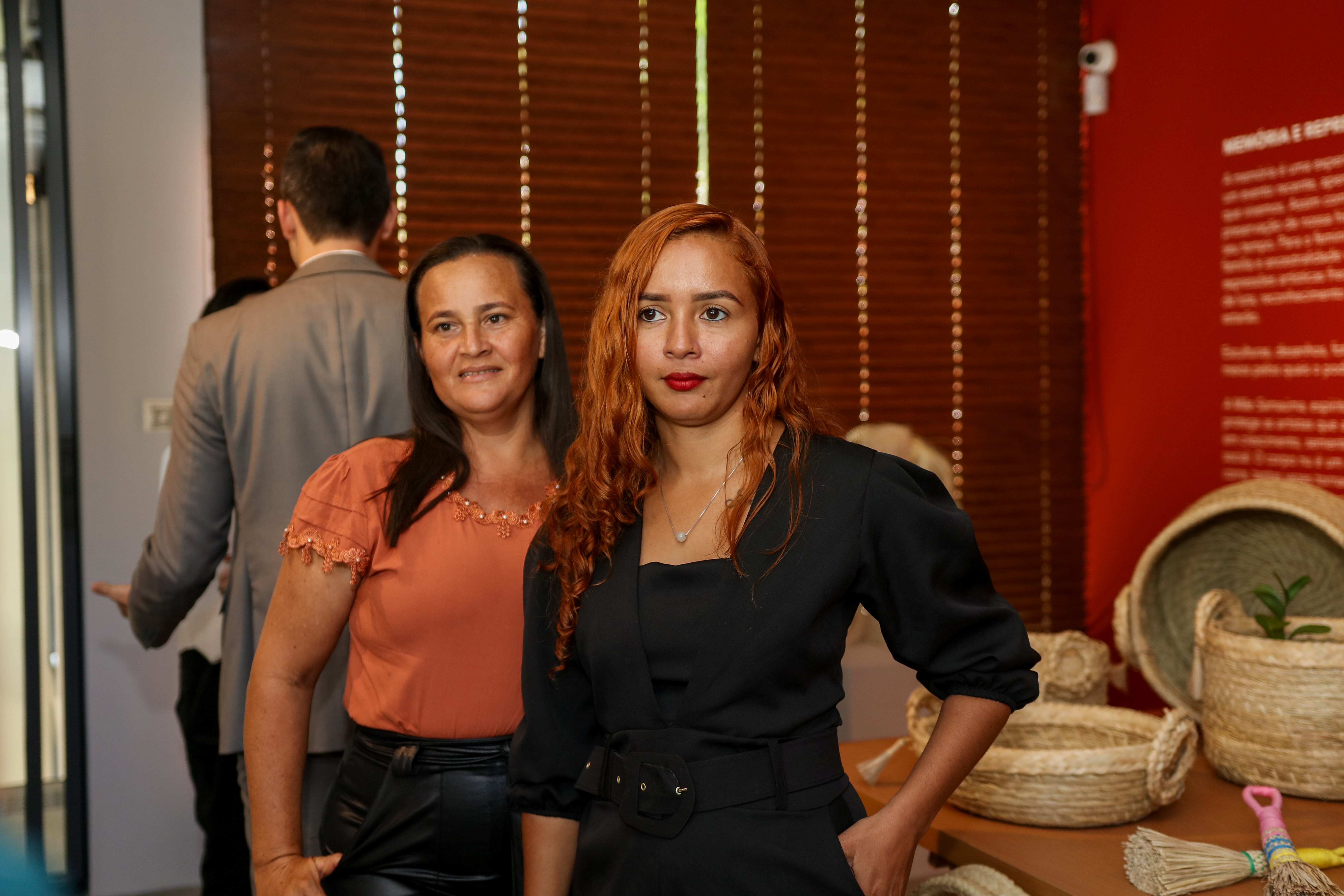 Fátima Laine Sousa Santos frente a una exposición de productos artesanales tejidos con fibra de carnauba en un evento en una sala. A su lado, otra participante sonríe. 