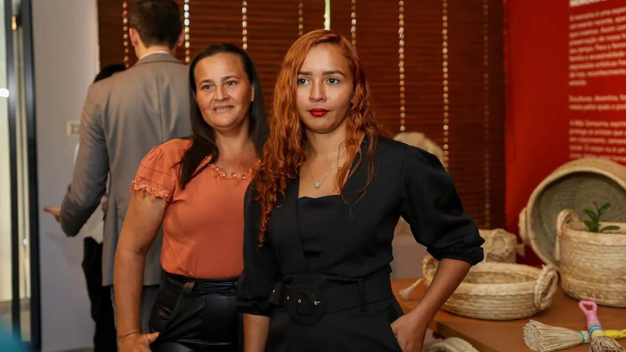 Fátima Laine Sousa Santos frente a una exposición de productos artesanales tejidos con fibra de carnauba en un evento en una sala. A su lado, otra participante sonríe.