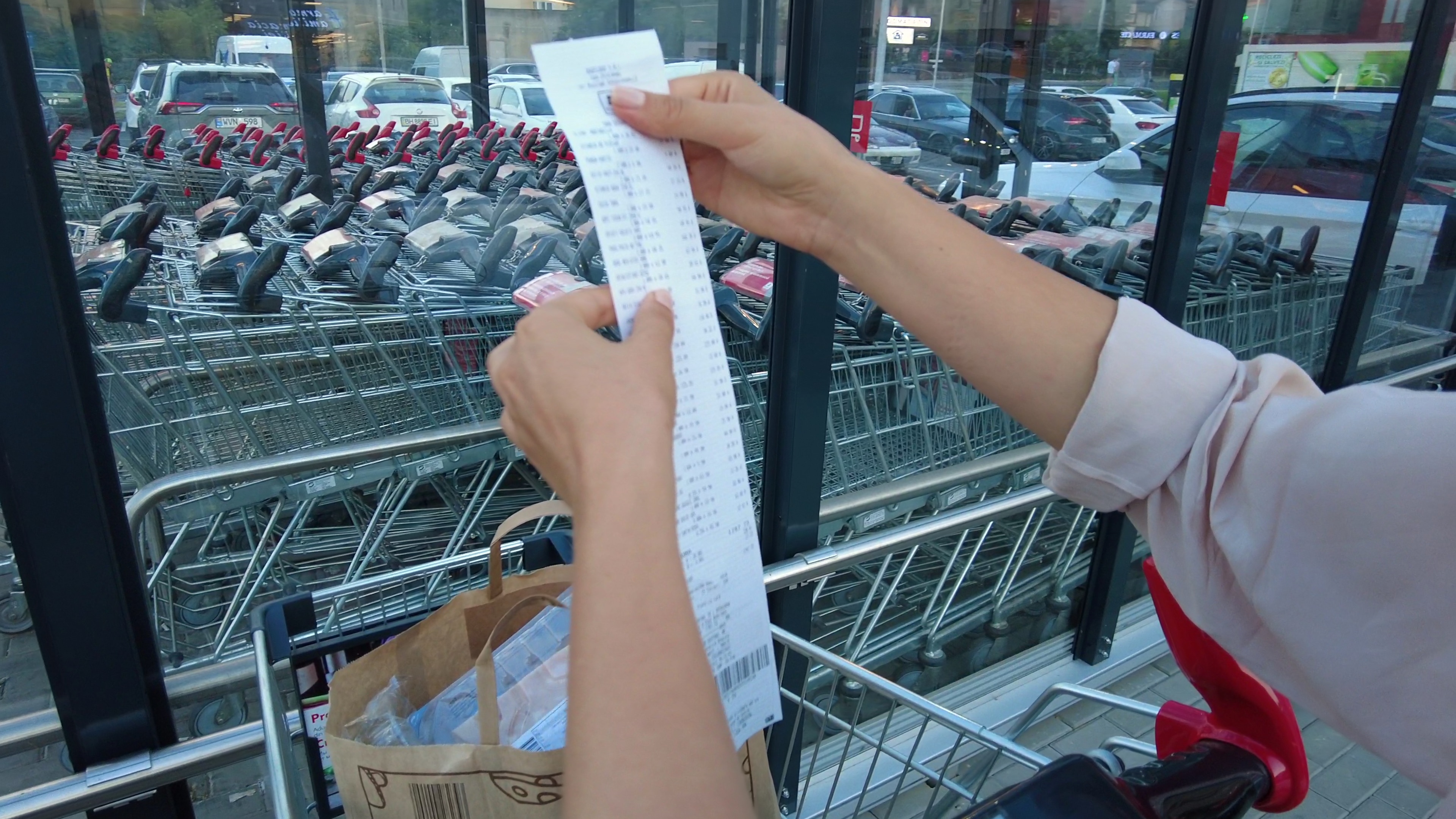Close-up of a woman's arms and hands as she holds a grocery receipt above a bag in a shopping cart. A row of shopping carts and a parking lot are in the background.