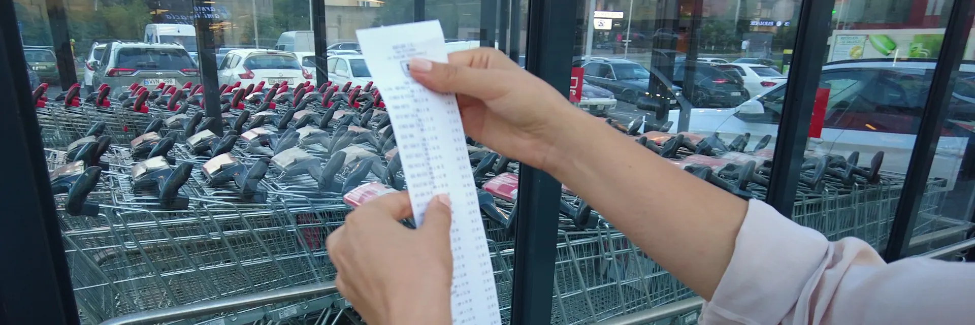 Close-up of a woman's arms and hands as she holds a grocery receipt above a bag in a shopping cart. A row of shopping carts and a parking lot are in the background.