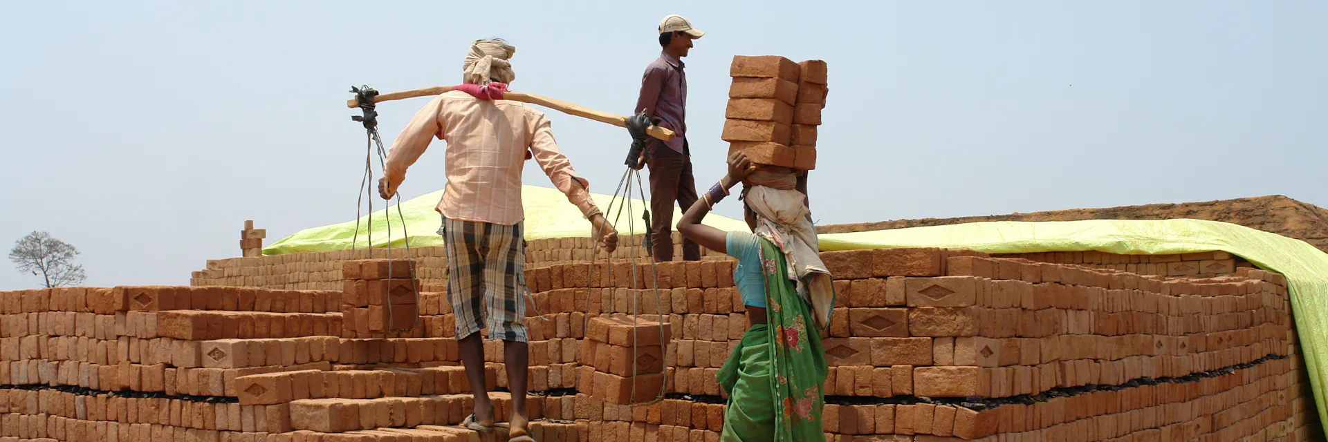 Two brick kiln workers in Andhra Pradesh, India, carrry big piles of bricks up a set of stairs made of bricks. The woman carries 10 bricks on her head. The man balances two piles of bricks which are tied to a long pole balanced across his shoulders. It is daytime. (2013)