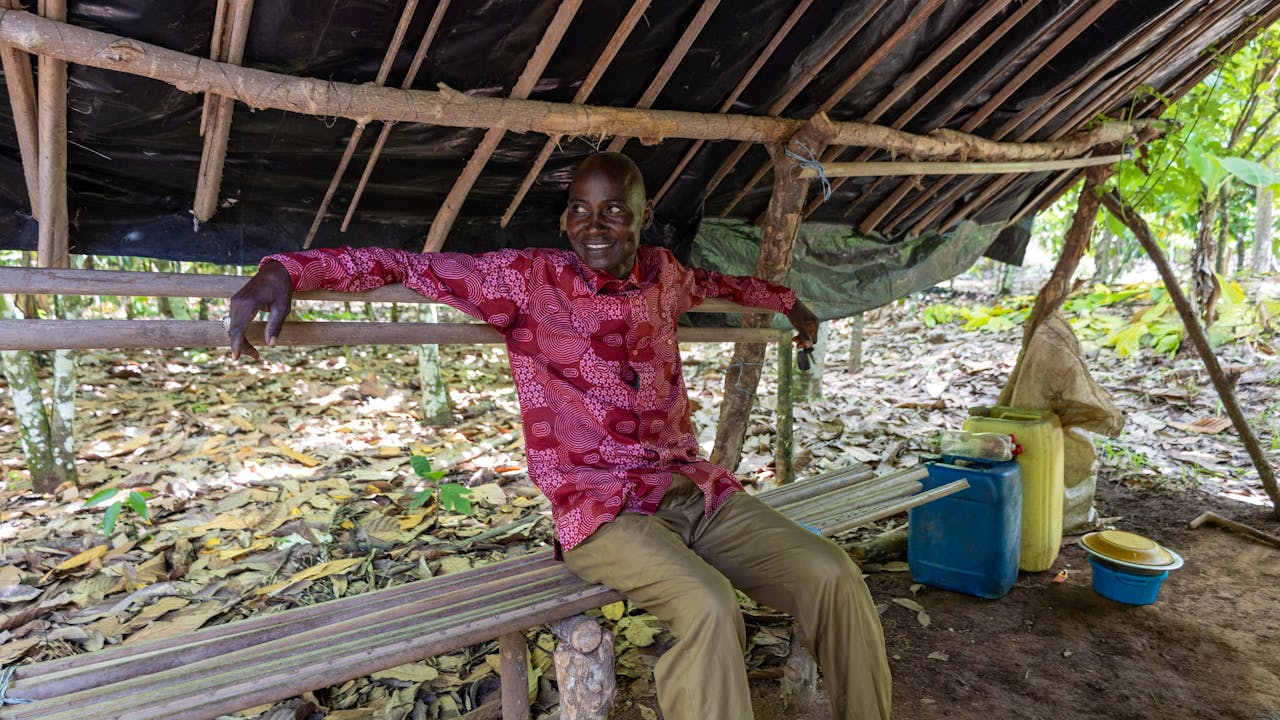 Yabao Oumarou está sentado en un banco bajo un techo de madera. Él sonríe apoyado a la estructura. (2024). 