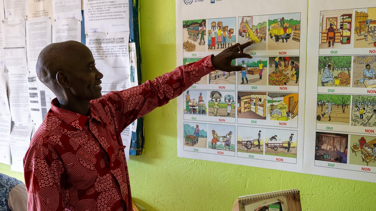 Yabao Oumarou señala los afiches expuestos en la pared. Los carteles muestran cómo los trabajadores de la plantación pueden mejorar la salud y la seguridad en el trabajo. (2024). 