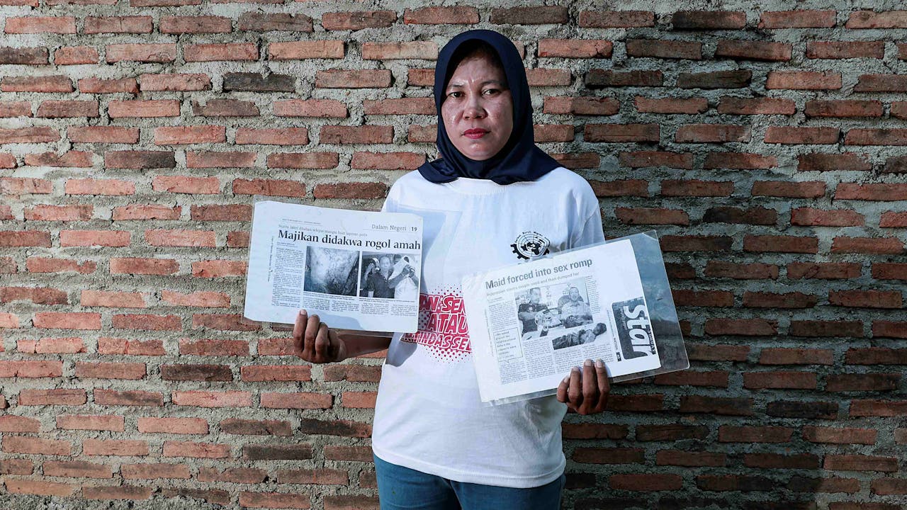 Win Faidah mira a la cámara y sostiene recortes de periódico sobre su experiencia. El titular de uno de ellos reza: "Asistente doméstica abusada sexualmente". Se sitúa de pie frente a un muro de ladrillos. 