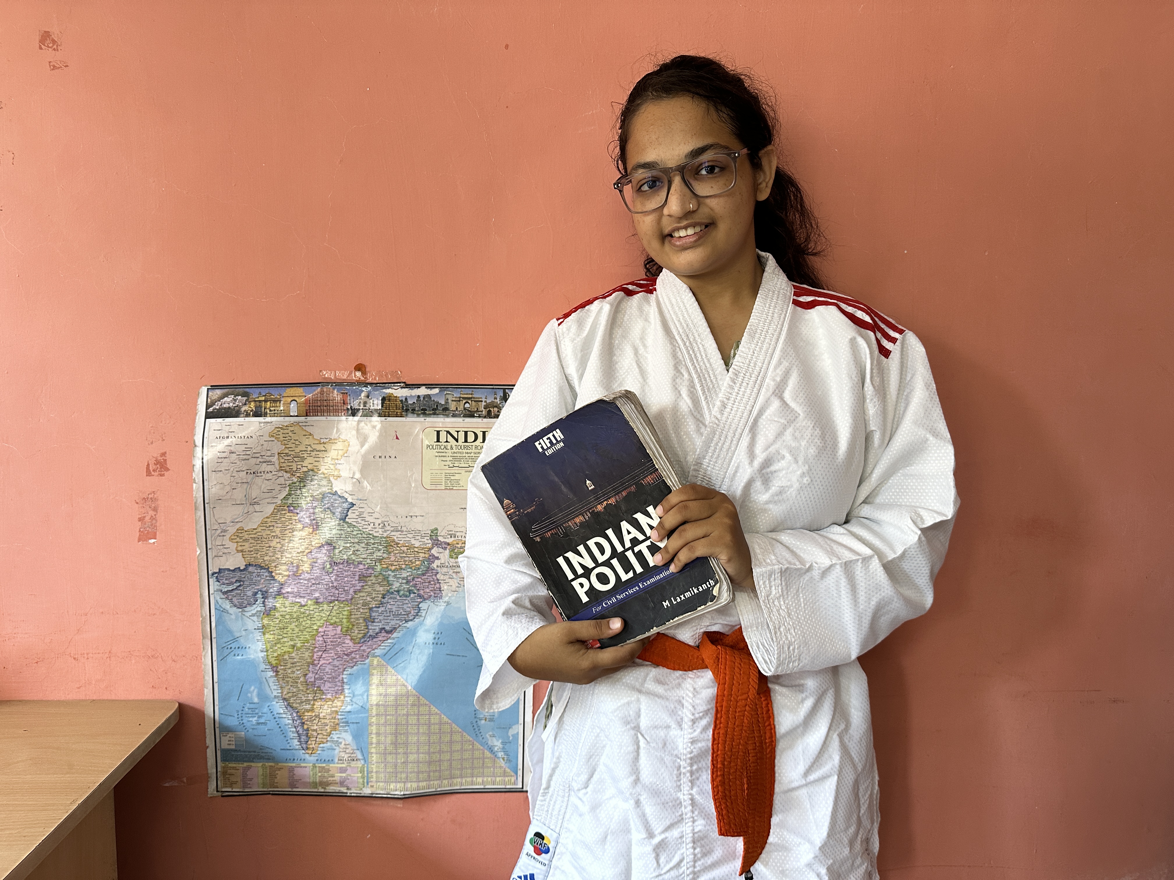 Reena est debout dans sa chambre, la carte de l’Inde affichée sur le mur derrière elle. Elle porte sa tenue de karaté et tient un livre sur la politique indienne dans les mains. Surat, Inde. (2024)