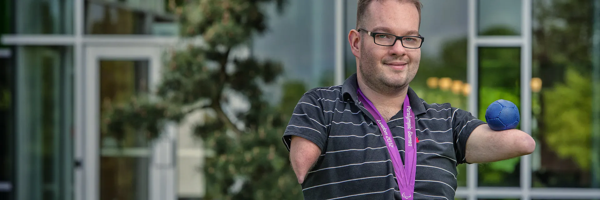 2012 Paralympic medallist Josh Vander Vies sits in front of glass building. He wears his 2012 Paralympic medal and holds a boccia ball. (2014)