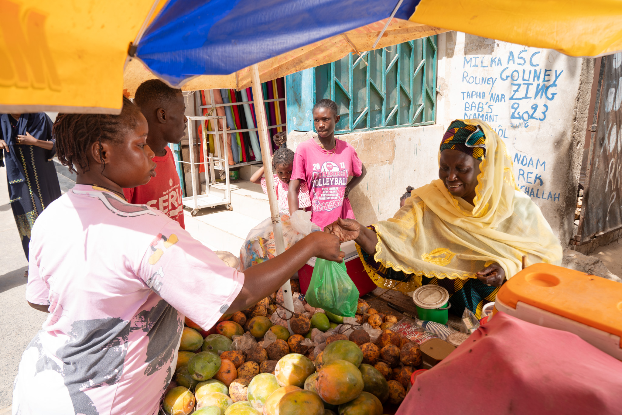 Astou Gningue sells fruit to a customer at a small stand in front of her home. Pikine, Senegal. (2024)