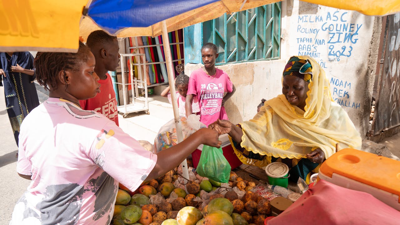 Astou Gningue sells fruit to a customer at a small stand in front of her home. Pikine, Senegal. (2024)