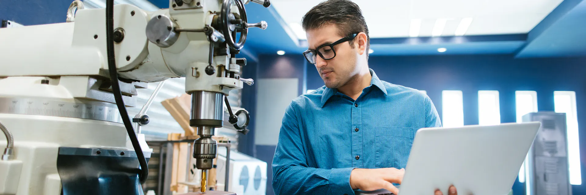 A young, male engineer works on calibrating a large drill in a workshop. A laptop rests on his left arm for reference.