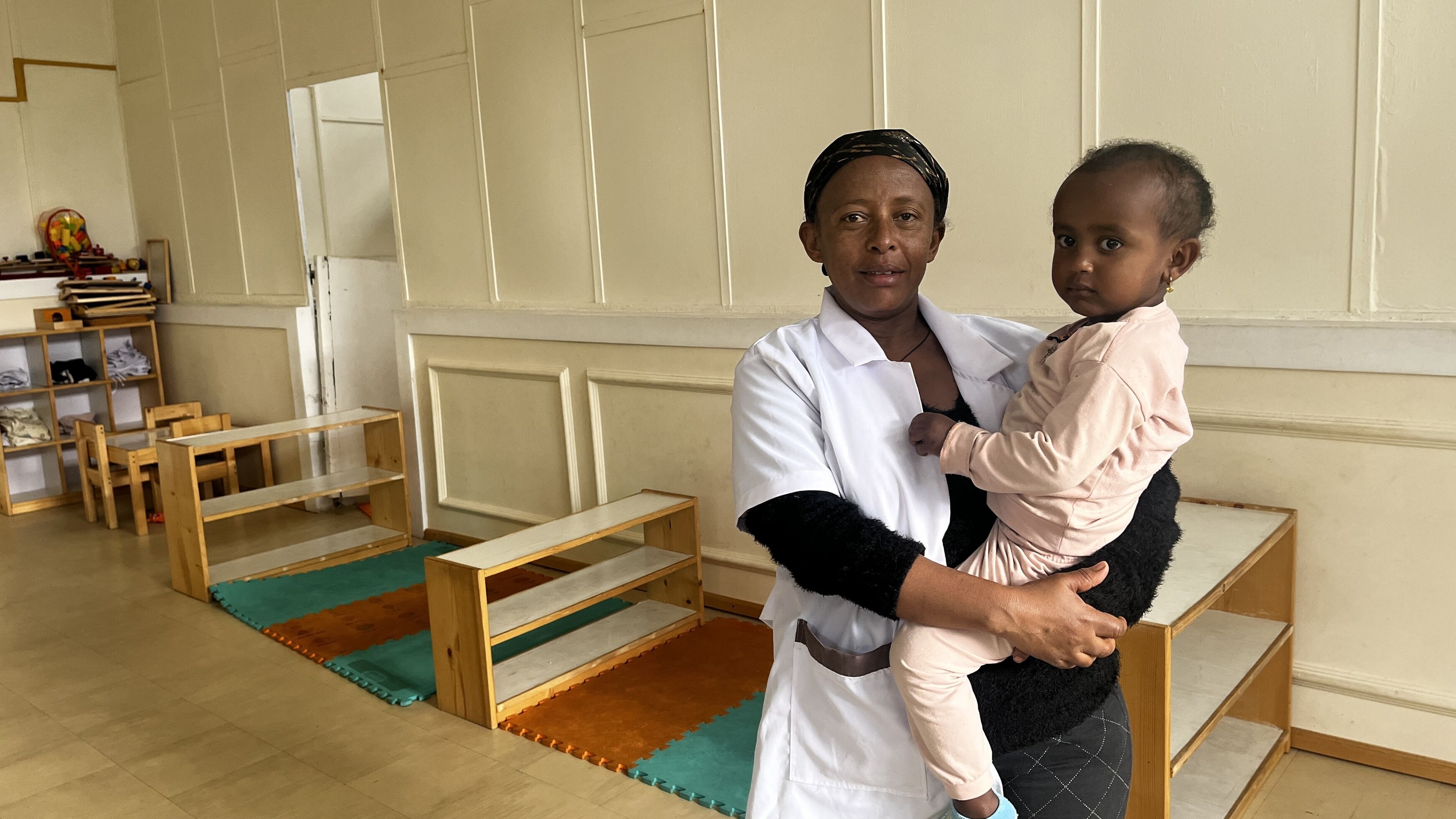 A trained childcare worker at the ILO-supported childcare centre holds Meseret Tamru's daughter. In the room, there are playmats, shelves and toys as well as furniture for small children. (Ethiopia, 2024)