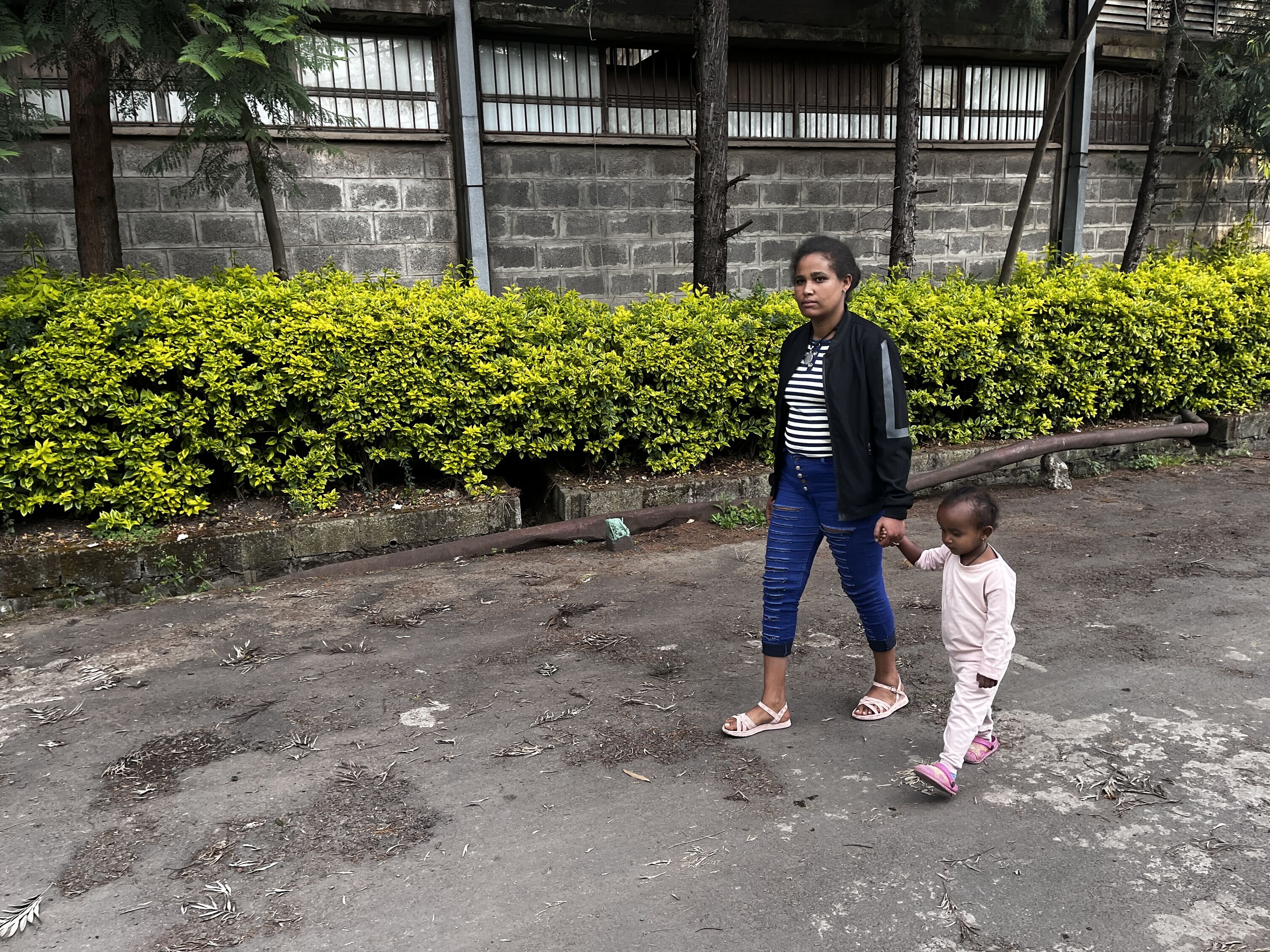 Meseret Tamru and her young daughter walk along a street in Addis Ababa, holding hands. Behind them, there is a building and greenery. (Ethiopia, 2024)