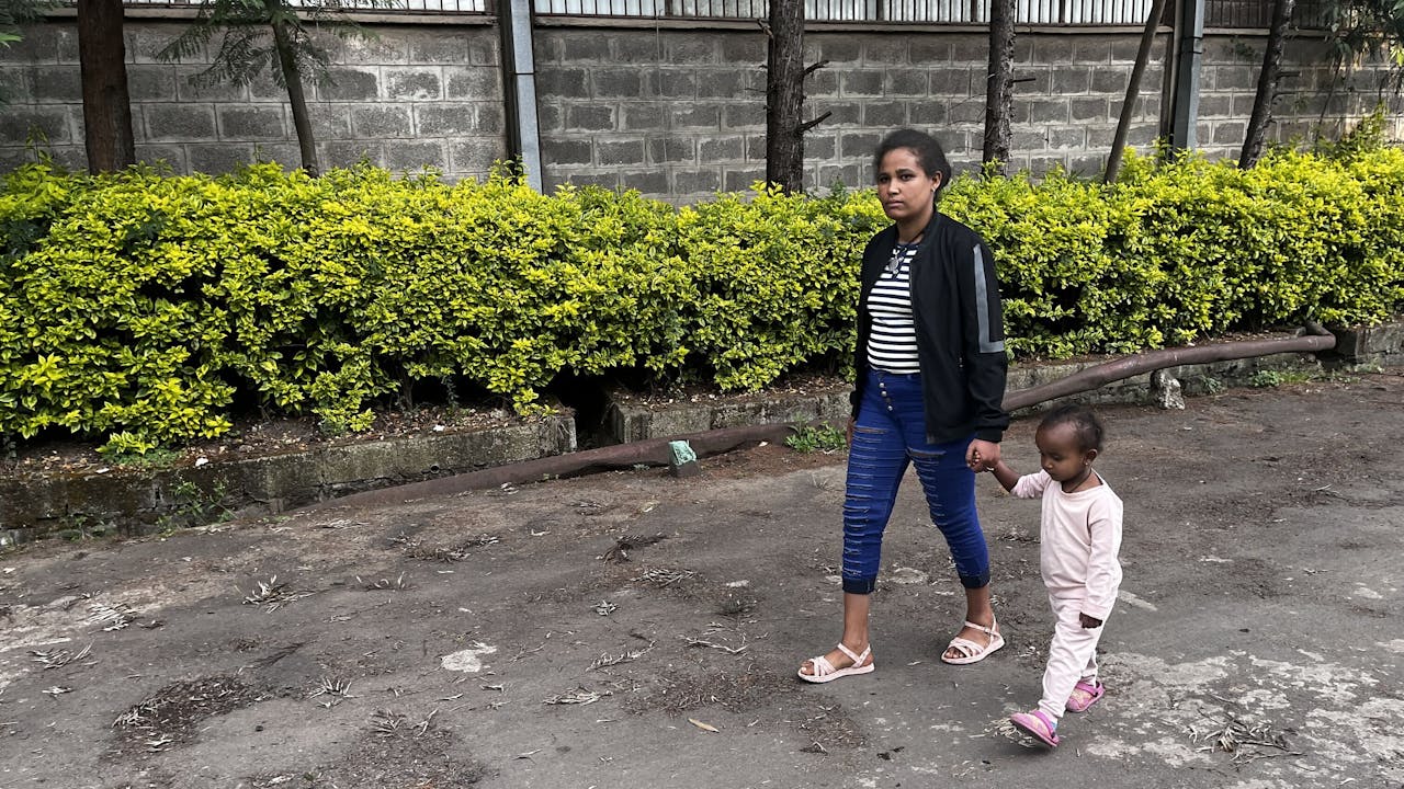 Meseret Tamru and her young daughter walk along a street in Addis Ababa, holding hands. Behind them, there is a building and greenery. (Ethiopia, 2024)