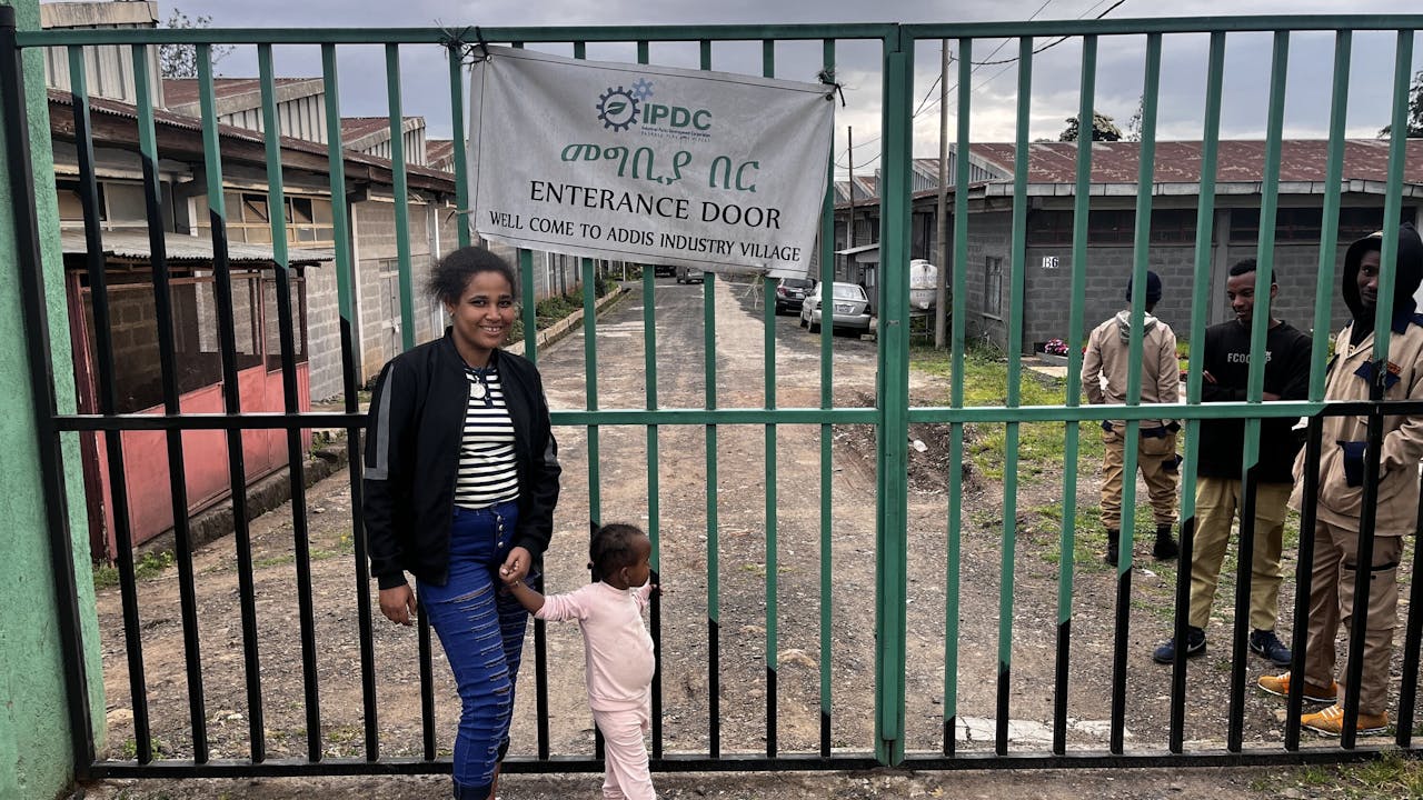 Meseret Tamru stands outsides the gates of the industrial park where she works. She smiles. Her young daughter holds her hand and looks through the gates. (Ethiopia, 2024)