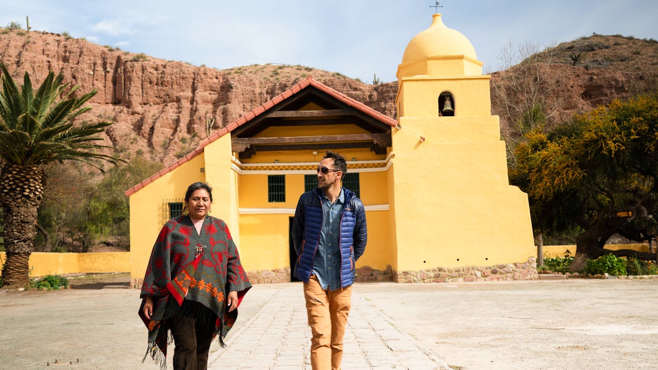 Celestina y un turista caminan frente a la iglesia de la aldea, pintada de amarillo.