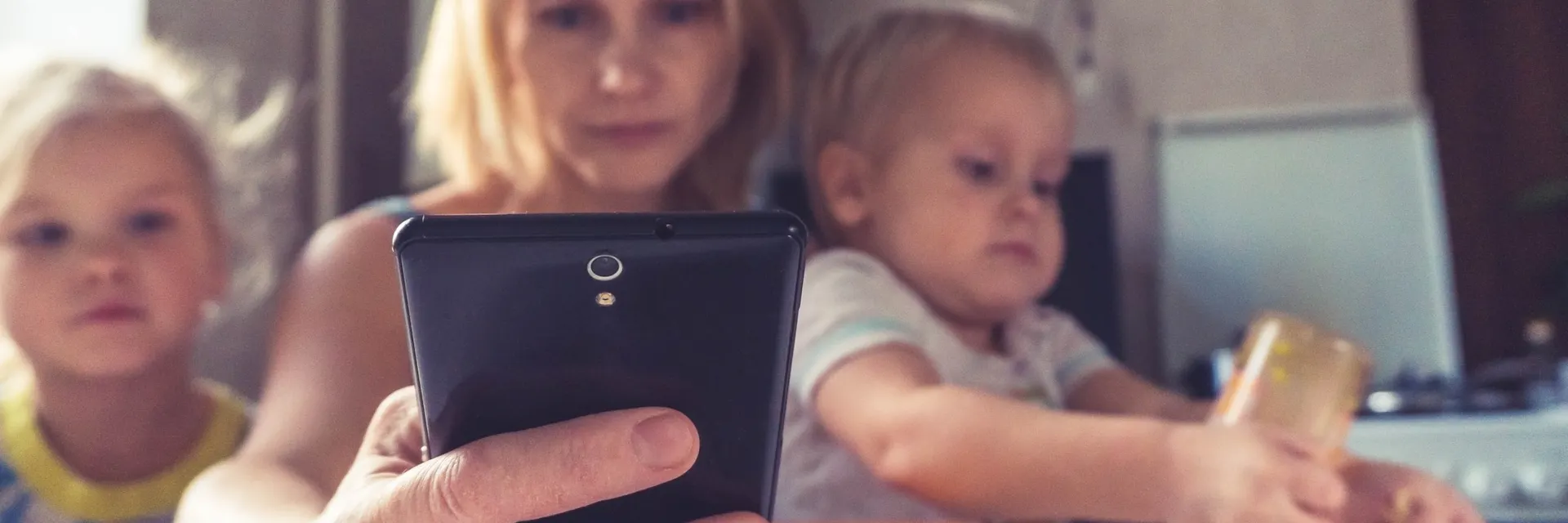 A working mother uses smartphone while sitting with her children at the kitchen table.