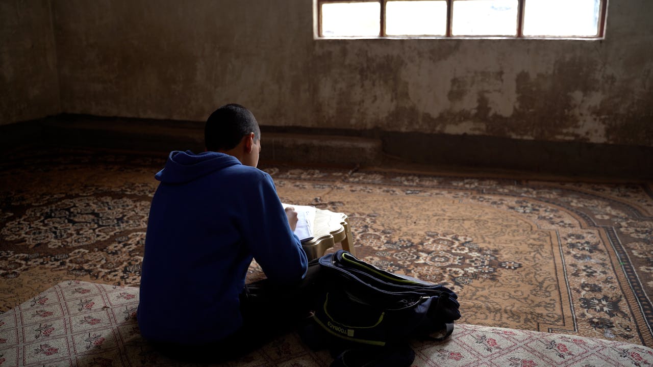 Akram est assis dans une pièce vide, meublée d'un tapis et d'une petite table, et fait ses devoirs.  