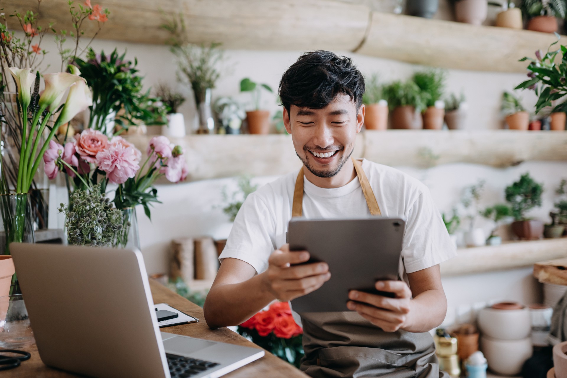 Asian male florist, owner in small legged florist shop, using digital tablet while working on laptop against flowers and plants.  