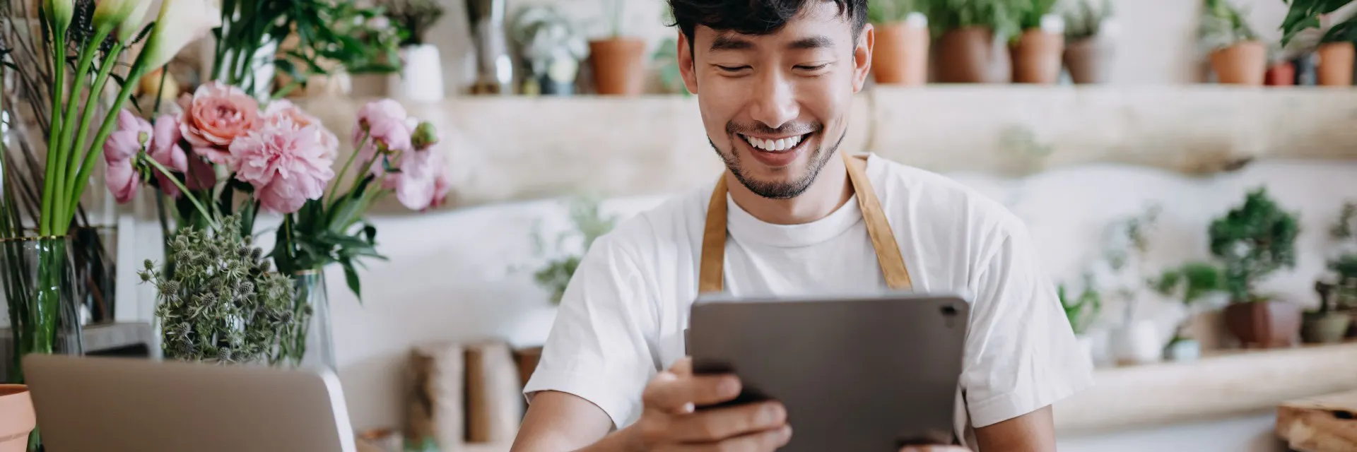 Asian male florist, owner in small legged florist shop, using digital tablet while working on laptop against flowers and plants.