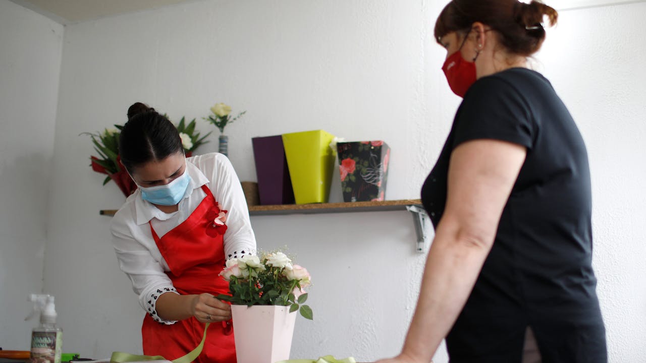 Mariam Kobalia prepares white roses in a vase for a customer.  Both Mariam and the customer wear a mask. 