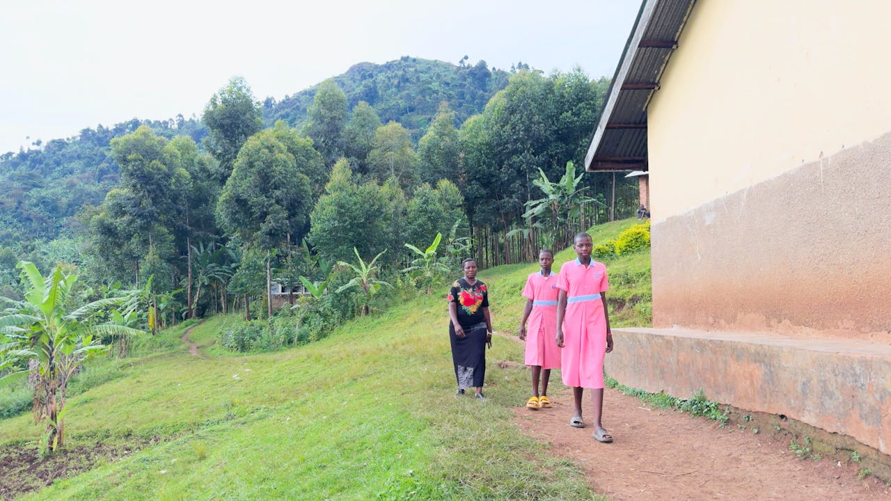 Rebecca Nakayega Wandegge camina por un sendero junto a un edificio en una zona rural. Sus dos hijas van por delante, vestidas con el uniforme escolar. (2025)