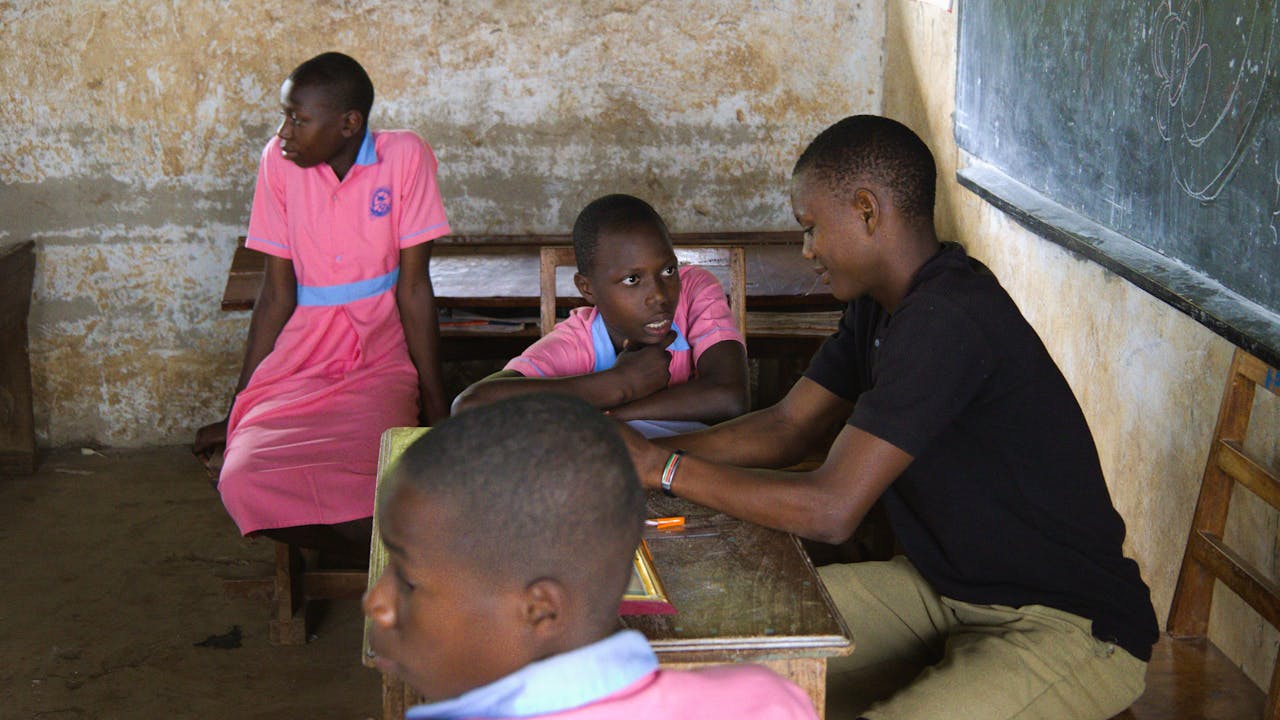Las dos hijas de Rebecca Nakayega Wandegge, sentadas en un aula con su maestro. (2025)