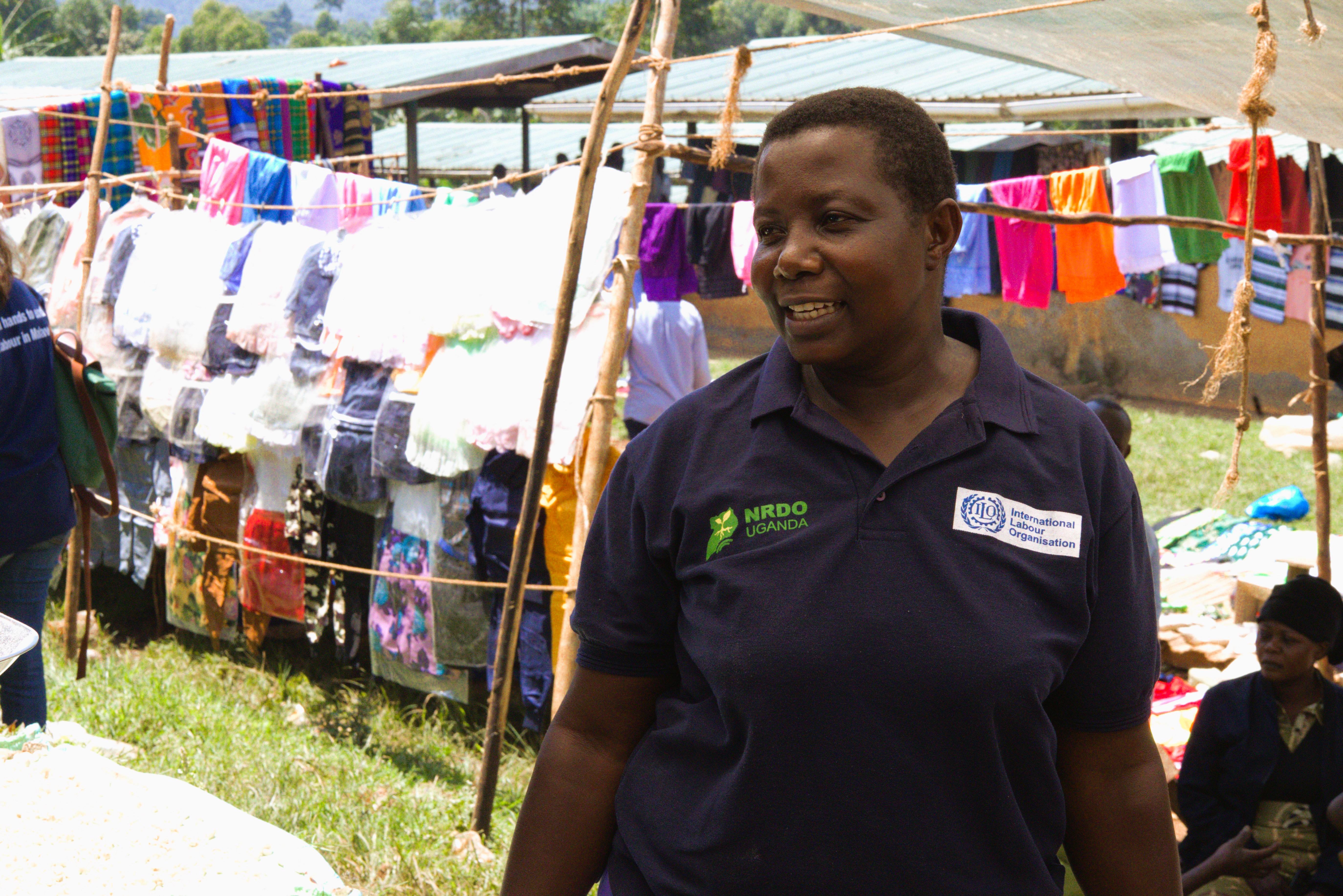 Rebecca Nakayega Wandegge stands in the market place and wears a t-shirt with the International Labour Organization (ILO) logo. She smiles. 