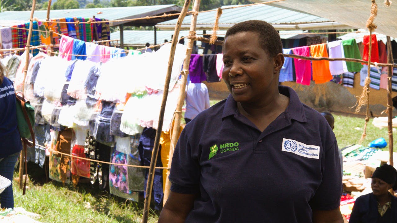 Rebecca Nakayega Wandegge de pie en el mercado, luciendo una camiseta con el logotipo de la Organización Internacional del Trabajo (OIT). Sonríe. (2025)