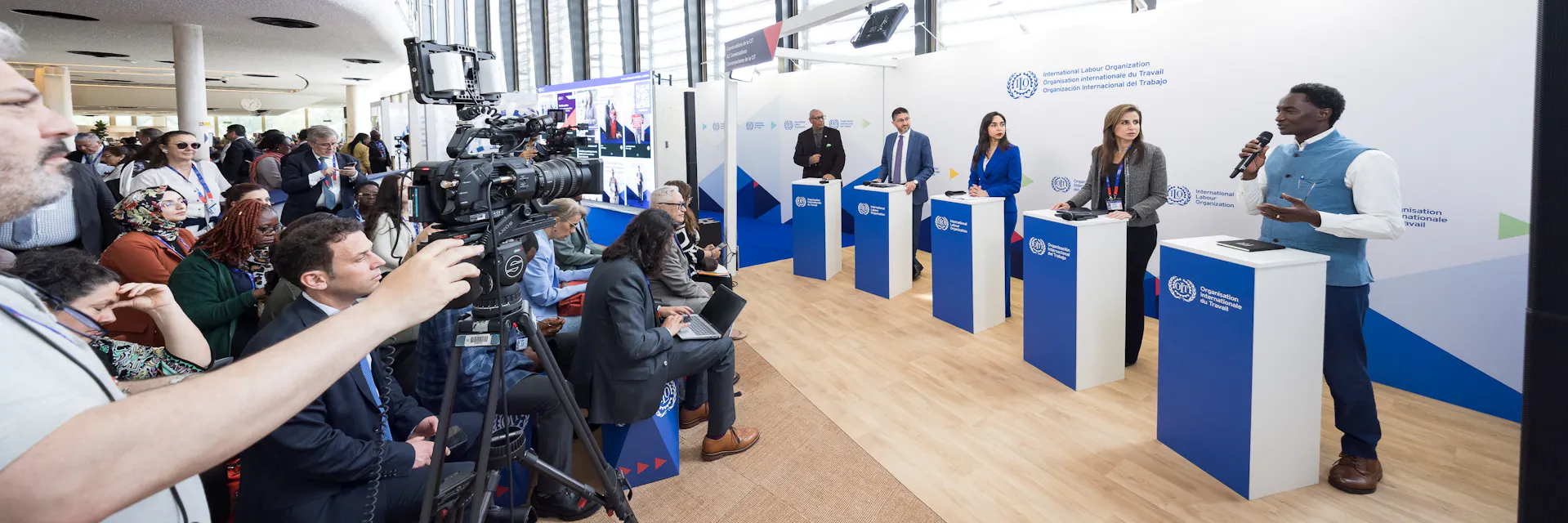 To the right are four panellists composed of two men and two woman and a male host. The panellist in the foreground holds a microphone and is speaking. To the left a crowd of people watch. In the foreground to the left, a male camera operator films the panellists.