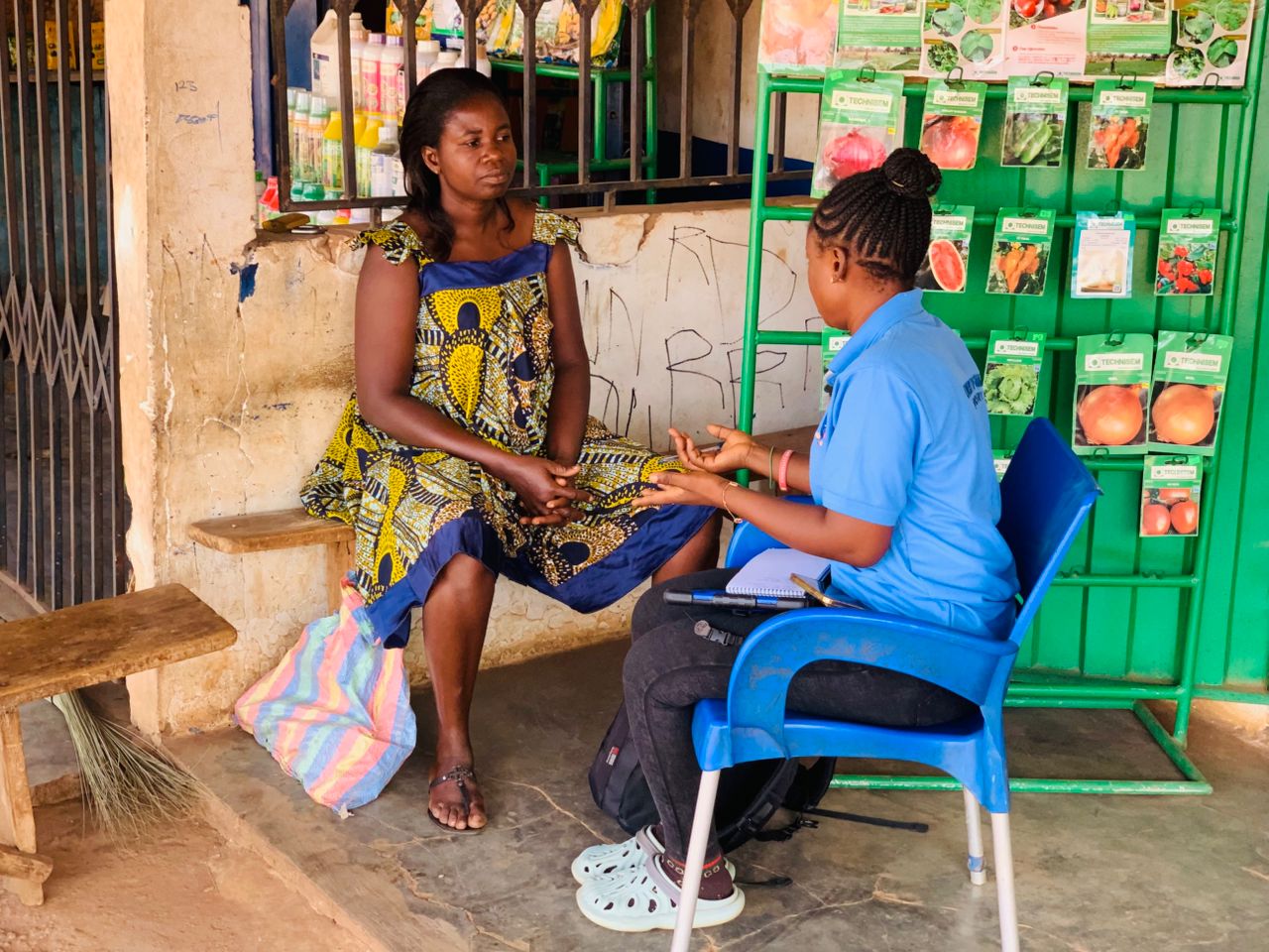 Kalian Sampoa Gumah conducts a survey with a female farmer. The farmer wears a brightly coloured dress.  The are seated outside a small grocery store next to a green metal stand with rows of seeds in packets. (Ghana, 2025) 
